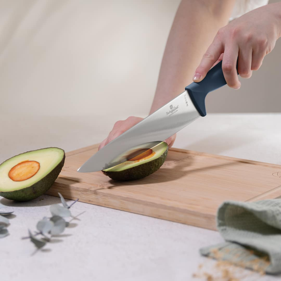 Person slices a ripe avocado in half on a wooden cutting board, removing the pit with a sharp chef's knife with a dark blue handle