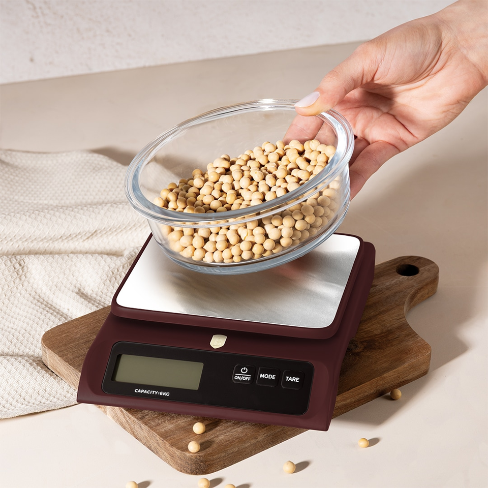 Hand placing a glass bowl of pale soybeans onto a digital kitchen scale on a wooden board