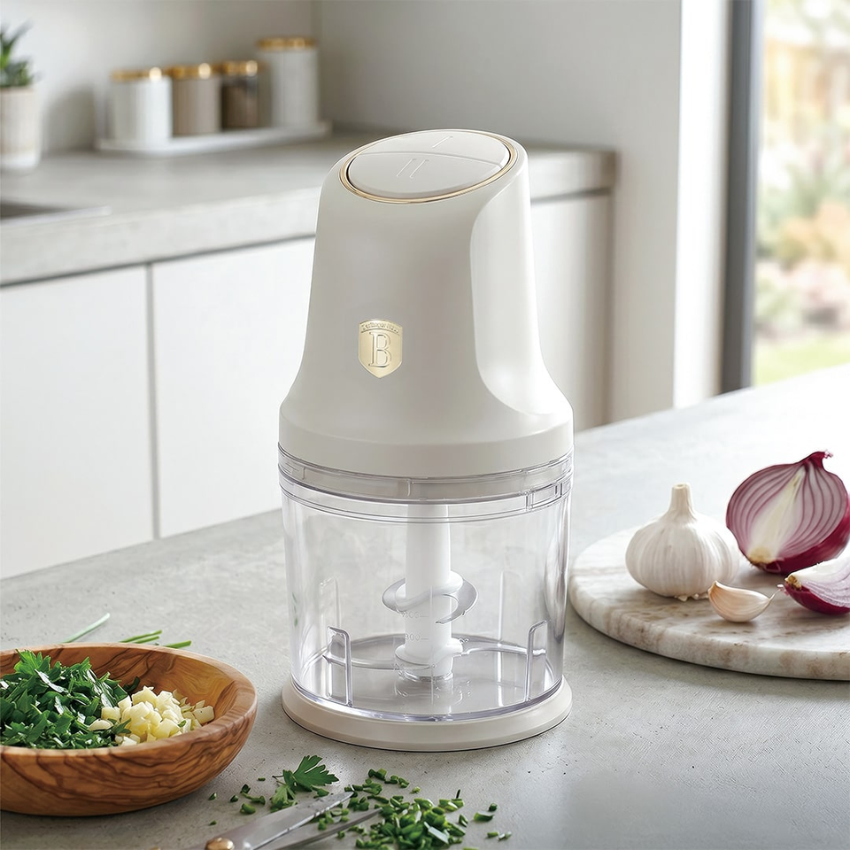 Compact white electric food chopper on a kitchen island with a clear bowl, blades visible inside, surrounded by chopped herbs and garlic.