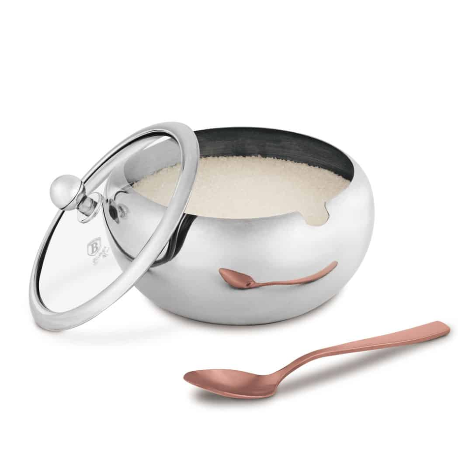 Polished stainless steel sugar bowl with its lid removed, filled with sugar, and a copper spoon beside it on a white background