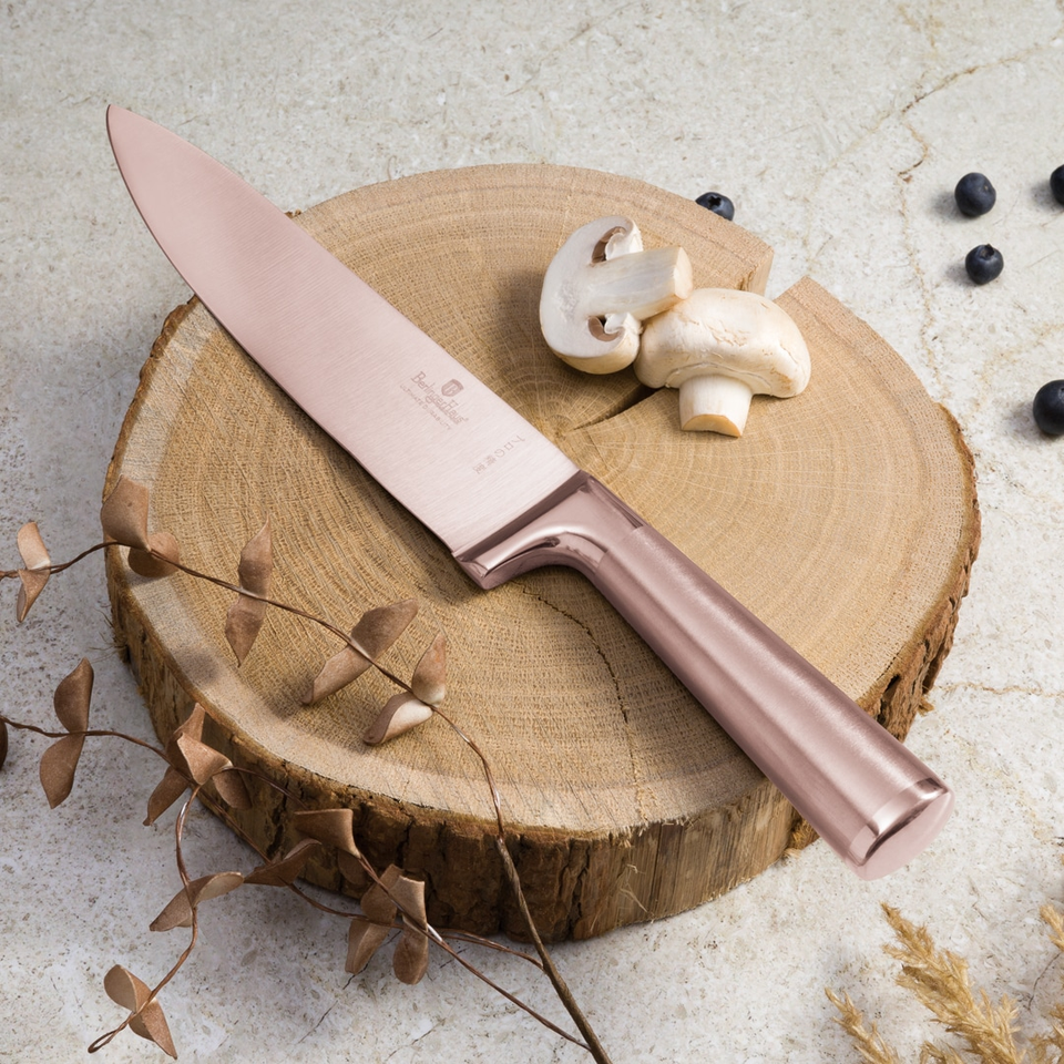 Rose-gold chef's knife lying across a tree-stump cutting surface with white mushrooms nearby and small berries in the background