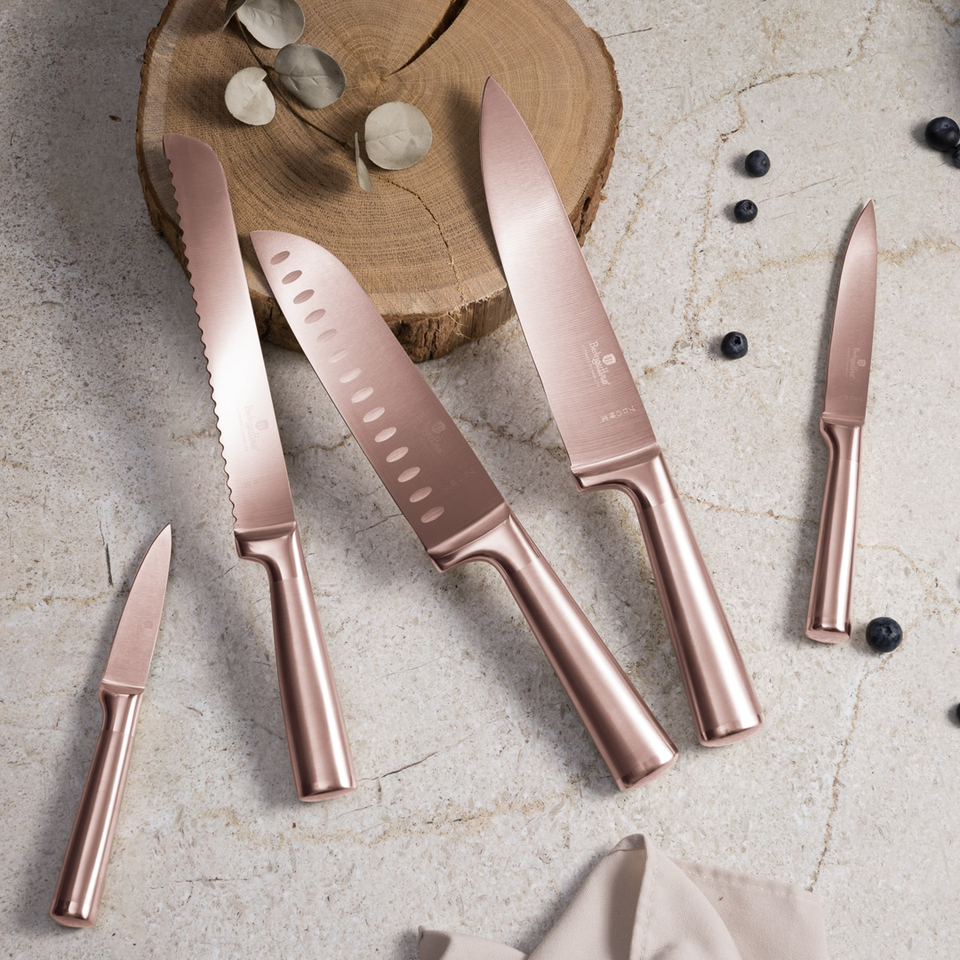 Rose-gold kitchen knife set laid out on a textured countertop with a wooden log slice and scattered berries in the background.