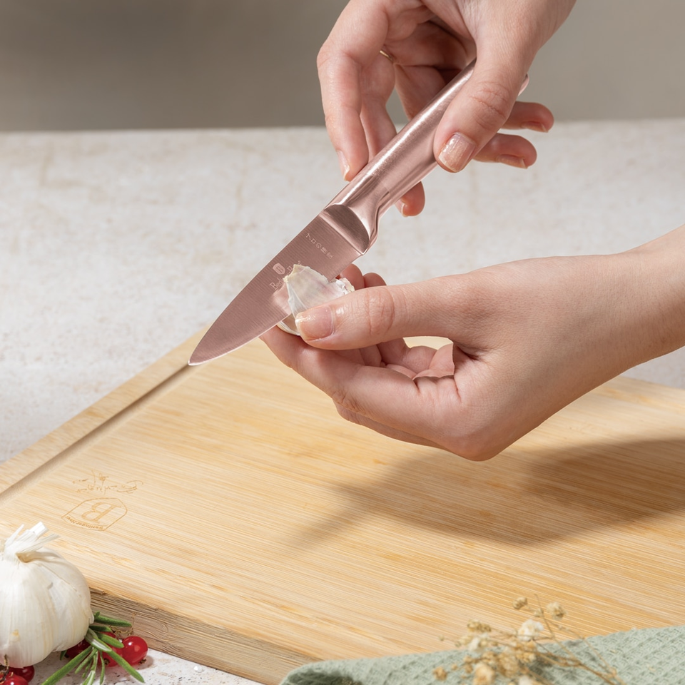 Person peeling a garlic clove with a small rose‑gold knife over a wooden cutting board, garlic head and sprigs nearby