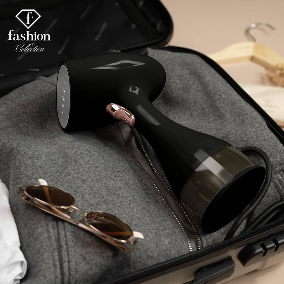 Black handheld garment steamer resting inside an open, gray-lined suitcase with sunglasses and clothing items nearby